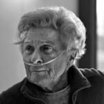 Black and white portrait of an elderly woman with a nasal cannula, expressing resilience and wisdom in her gaze.